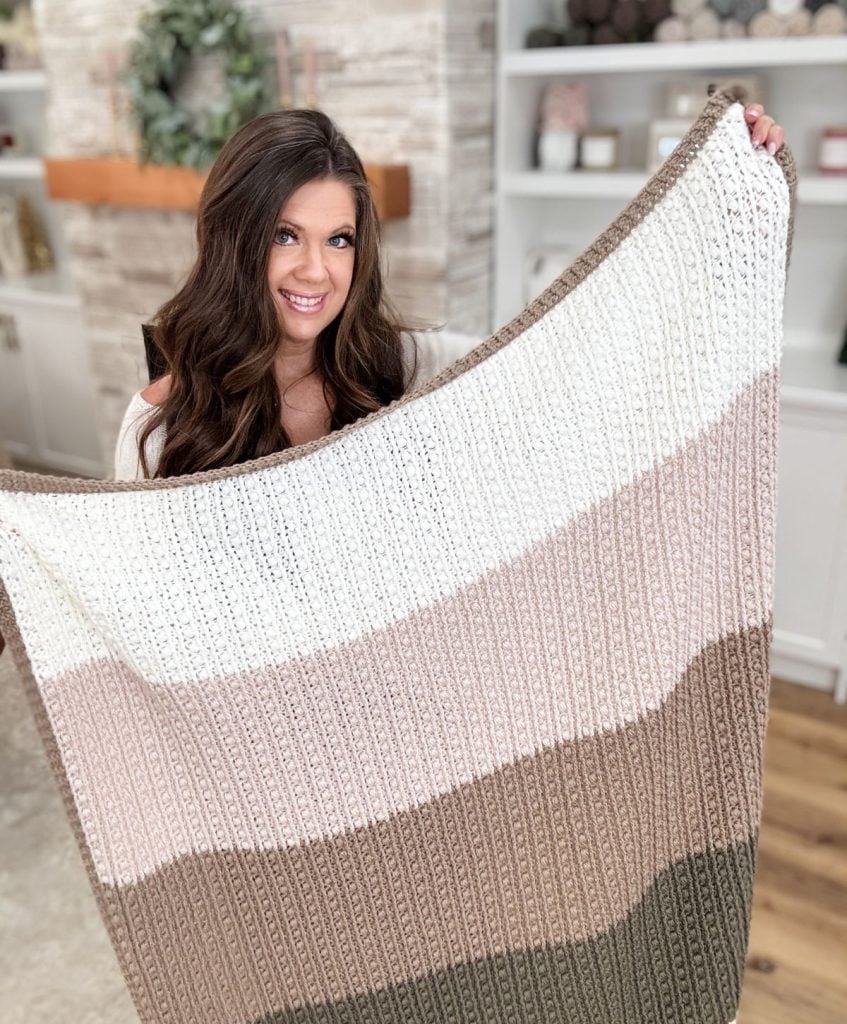 A person cradles a large crochet throw with horizontal stripes in shades of cream, beige, and brown, standing gracefully in a room adorned with shelves and a stone mantel in the background.