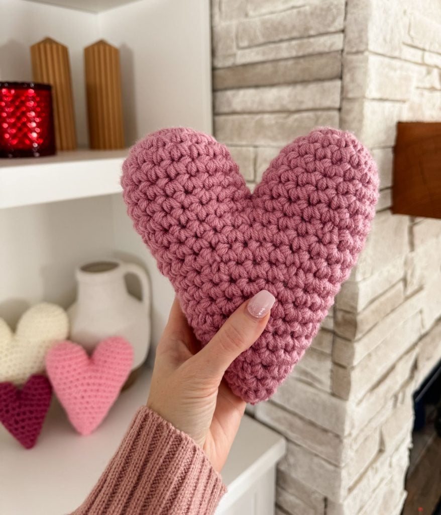 A hand holds a crochet heart in pink, showcasing intricate craftsmanship against a shelf adorned with decorative items, including more crocheted hearts and a red candle.