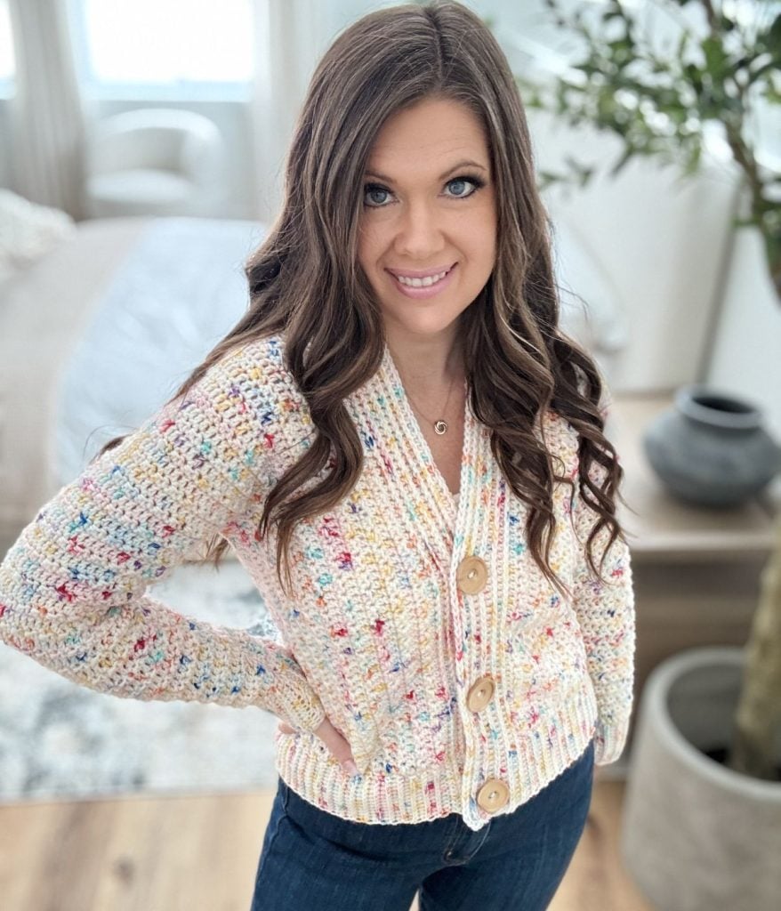 A person with long brown hair is smiling, wearing a multicolored Snowy Season Cardigan with large buttons and jeans, standing indoors.
