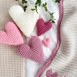 Crochet hearts in various shades of pink and white are displayed on a textured blanket with a leafy branch above.
