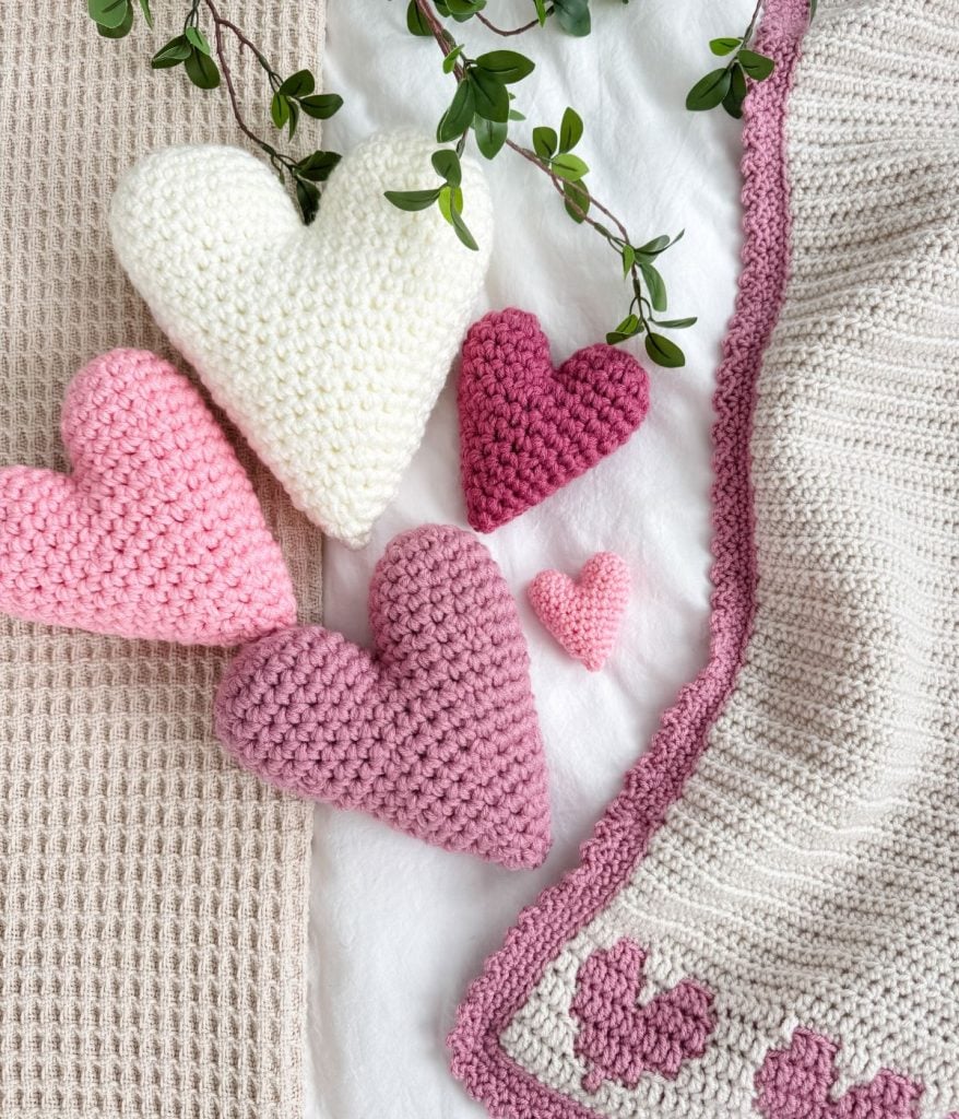 Crochet hearts in various shades of pink and white are displayed on a textured blanket with a leafy branch above.