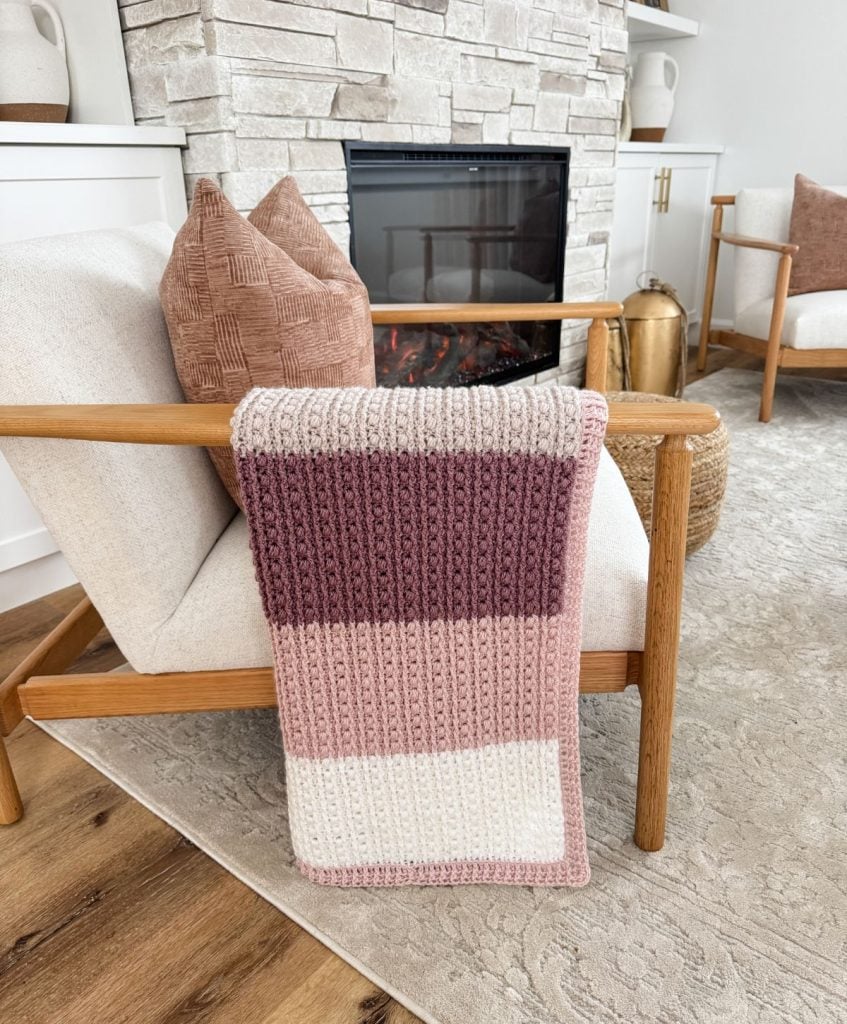 A crochet throw with pink, purple, and white stripes draped over a wooden armchair near a stone fireplace in a cozy living room.