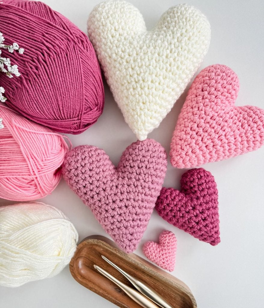 Crochet heart shapes in shades of pink and white are arranged with matching yarn balls and crochet hooks on a light surface, creating a harmonious display.