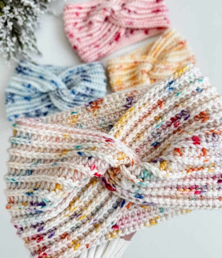 Four easy headbands with knot designs in pink, blue, yellow, and multicolored patterns are artfully arranged on a white surface alongside a sprig of greenery.