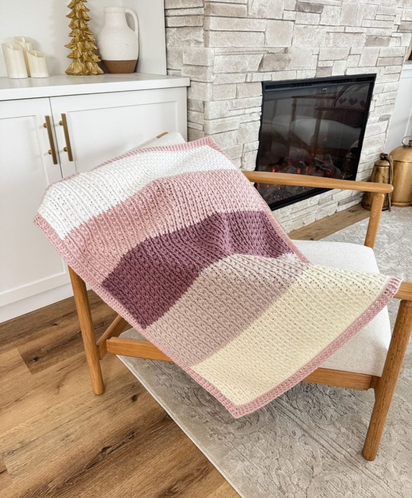 A cozy crochet throw with pink, purple, and white stripes is draped over a wooden chair. A stone fireplace and cabinet are in the background.