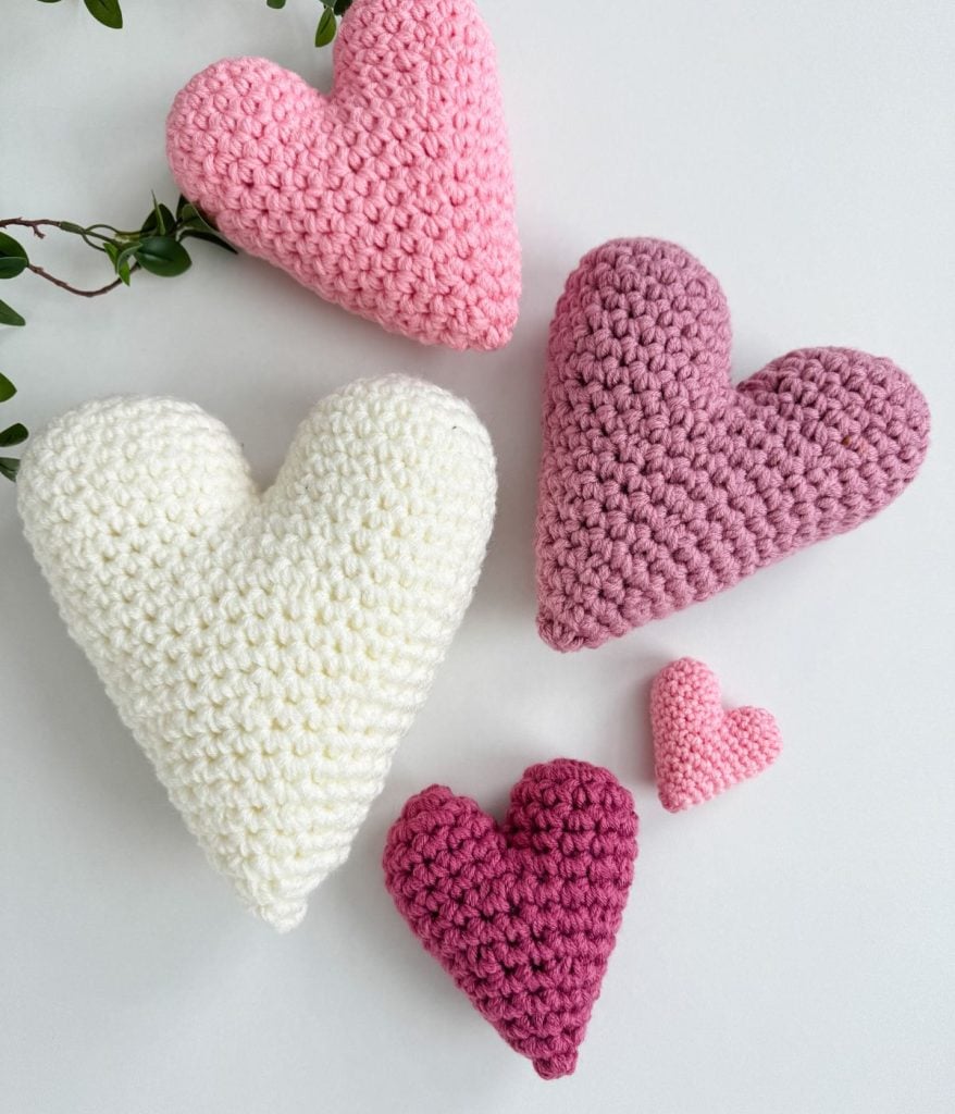 Five crochet hearts in varying sizes and shades of white, pink, and purple rest elegantly on a plain background with a sprig of green leaves.