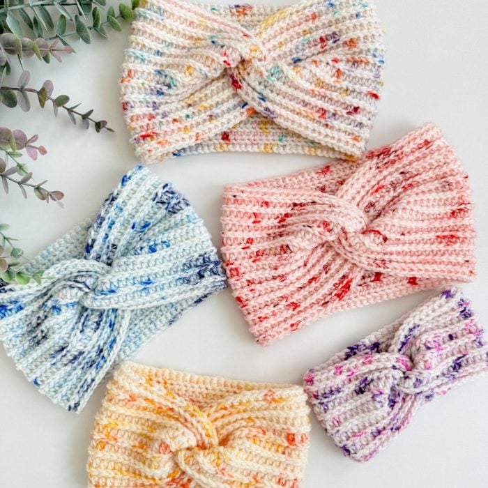 Five knitted easy headbands in various pastel colors with speckled patterns are arranged on a white surface. A green plant graces the upper left corner, adding a touch of nature.