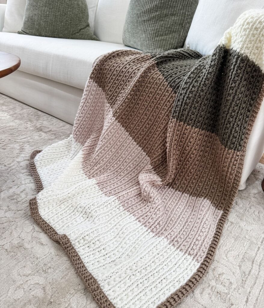A crochet throw with wide beige, cream, and brown stripes is draped over the arm of a white sofa with green textured cushions.