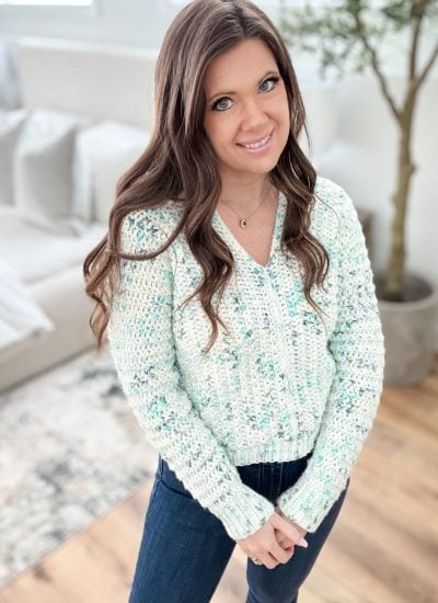 A person with long hair stands indoors wearing a light blue textured snowy season sweater and dark jeans, with a sofa and plant in the background.