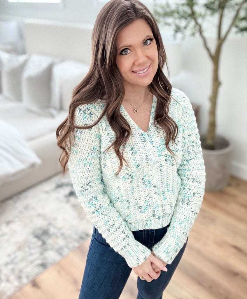 A person with long hair stands indoors wearing a light blue textured snowy season sweater and dark jeans, with a sofa and plant in the background.