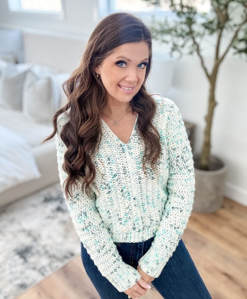 A woman with long hair stands indoors wearing a blue and white knit sweater reminiscent of a Snowy Season Sweater, paired with jeans and a necklace. In the background, a bed and plant complete the cozy scene.