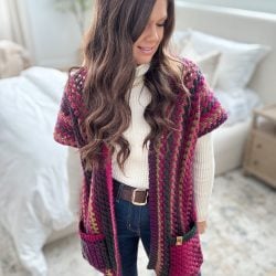 A person in a white turtleneck and jeans wears a multicolored knit vest with pockets, reminiscent of a Granny Pop Ruana design, standing in a room with a bed and nightstand.