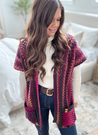 A person in a white turtleneck and jeans wears a multicolored knit vest with pockets, reminiscent of a Granny Pop Ruana design, standing in a room with a bed and nightstand.