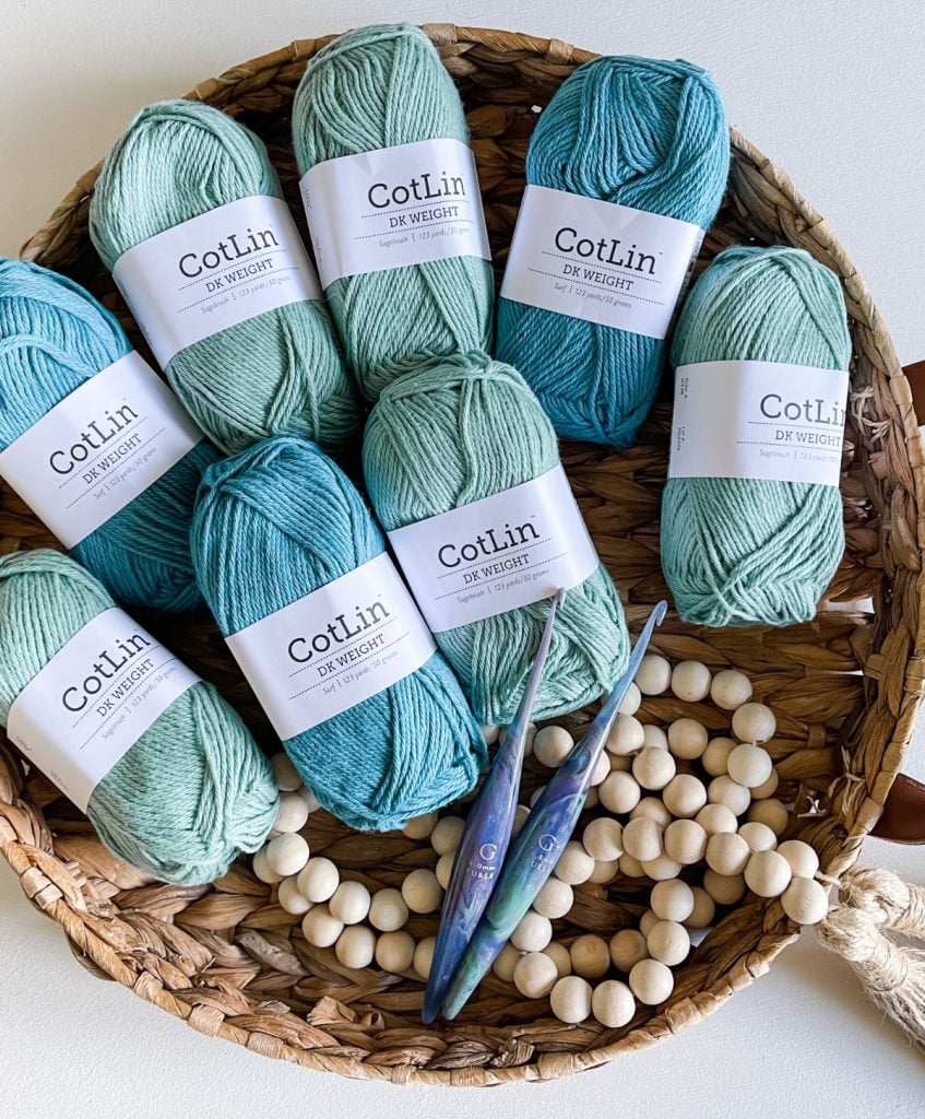 A cozy basket holds eight skeins of turquoise CotLin yarn, perfect for crafting a beautiful ruana. Nestled alongside are two crochet hooks and wooden beads, all resting on a woven mat.