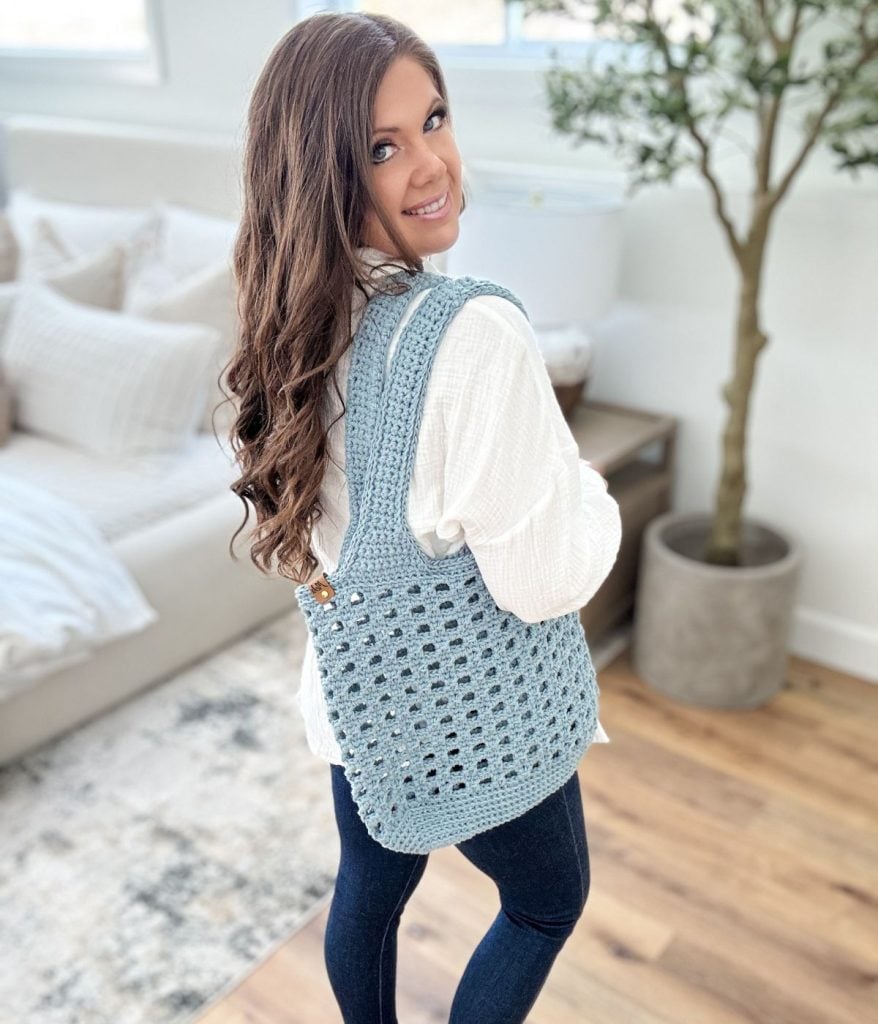 A person with long hair wearing a white shirt and blue jeans stands indoors, showing off a blue crochet mesh bag. A potted plant and a sofa create a cozy backdrop.