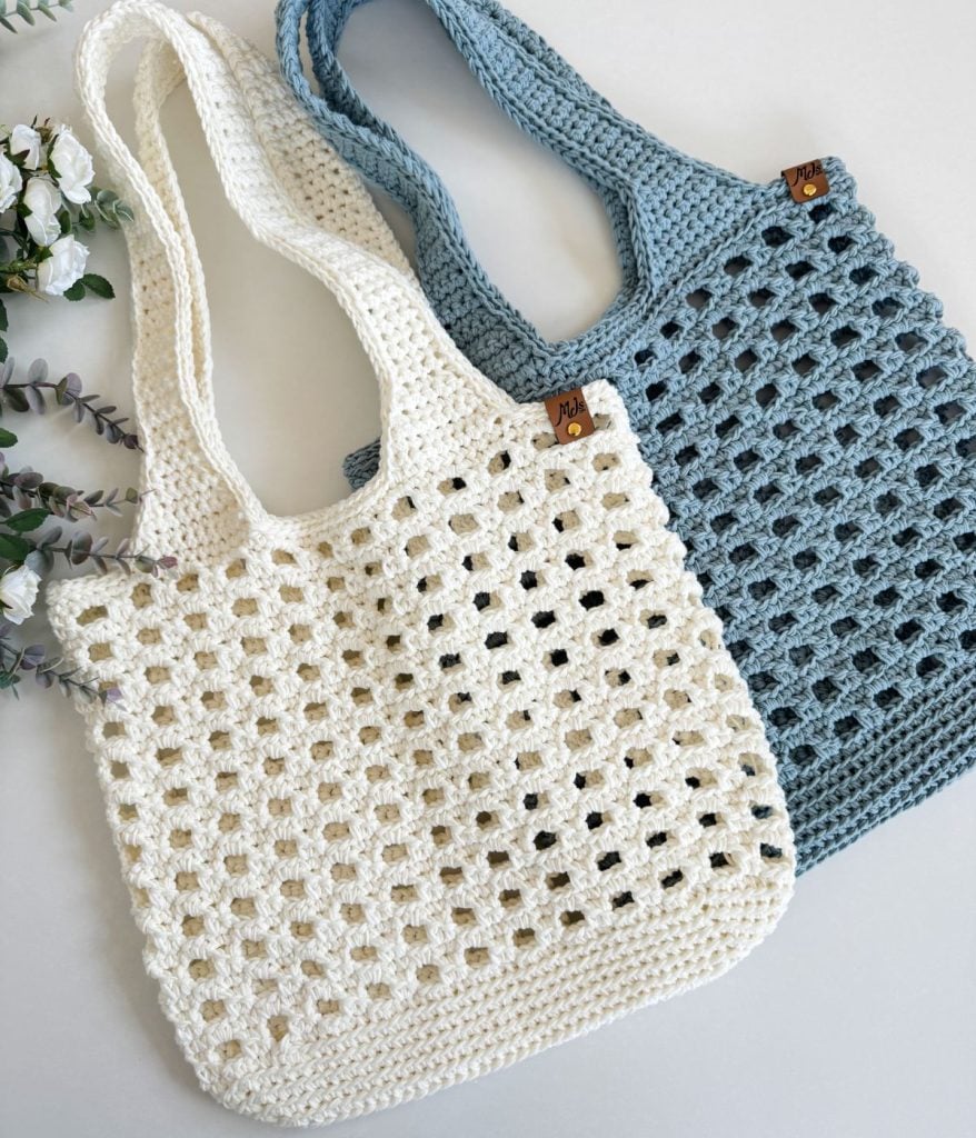 Two crochet mesh bags, one white and one blue with a grid pattern, feature wooden tags and are artfully arranged on a white surface next to decorative flowers.