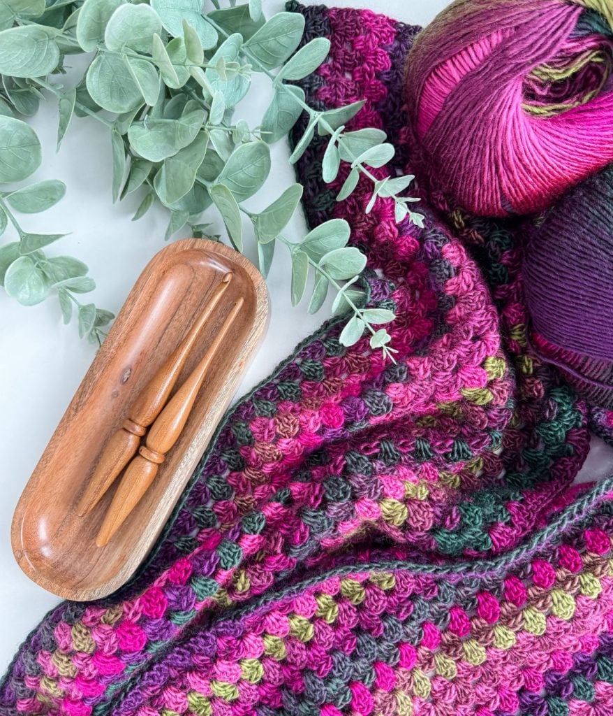 Colorful crochet blanket with magenta and green hues beside two wooden crochet hooks in a tray, a leafy branch, and multicolored yarn balls. Perfect for anyone looking for an easy crochet project or inspired by the vibrant Granny Pop Ruana!.
