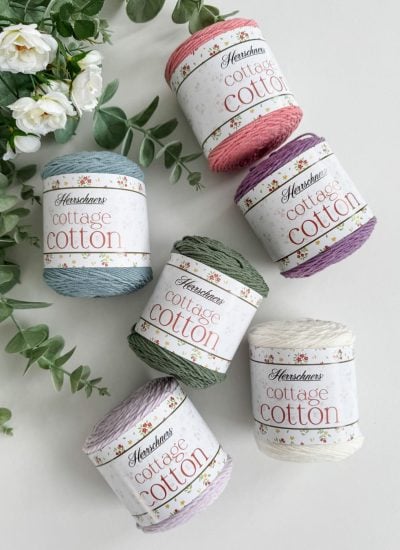 Six spools of Herrschners Cottage Cotton yarn in various colors, perfect for crafting a delicate crochet mesh bag, arranged on a white surface near green foliage.