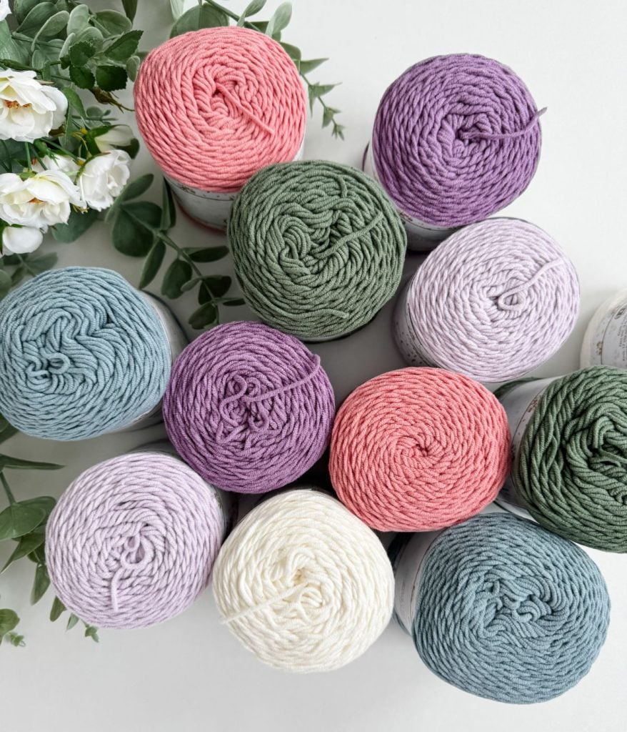 Twelve skeins of yarn in pastel purple, pink, green, and cream are arranged in a grid, ready to craft a charming crochet mesh bag. White flowers and green leaves elegantly decorate the edges.