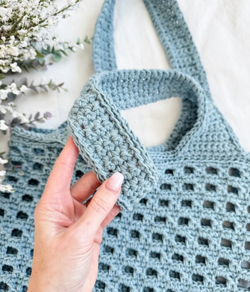A hand holds a small, rectangular, light blue crocheted piece above a matching crochet mesh bag adorned with a grid pattern. White and green flowers peek from the corner.