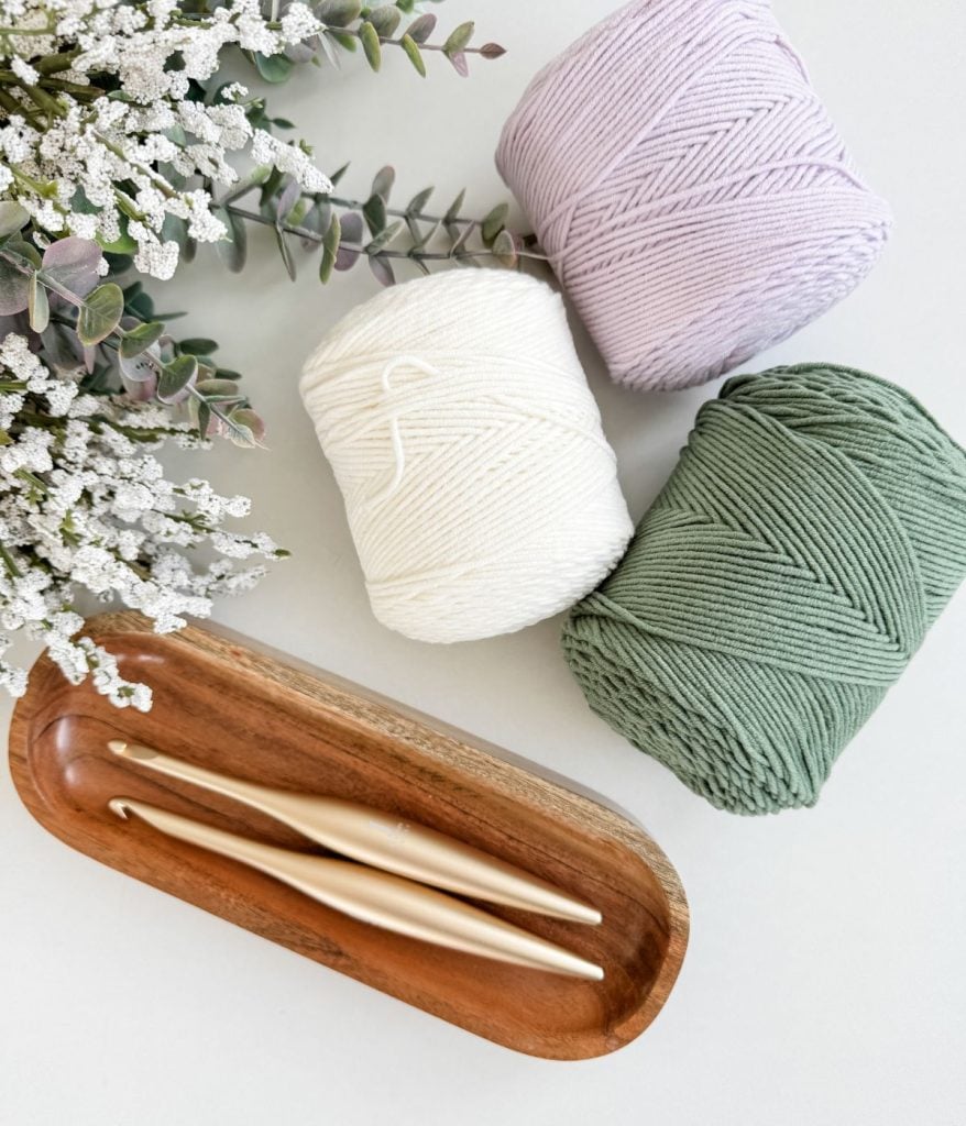 Three yarn balls in lavender, white, and green sit beside a wooden tray holding two crochet hooks, perfect for crafting a crochet mesh bag. White flowers and greenery grace the top left corner on a light surface.