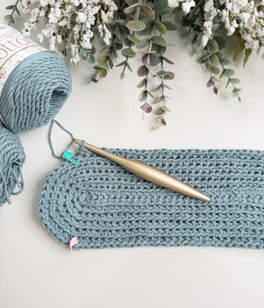 A crochet mesh bag is taking shape with blue yarn and a silver hook on a white surface, while lush greenery adds a touch of nature from above.
