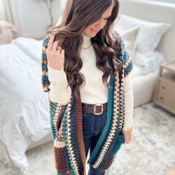 Person wearing a striped crochet cardigan, reminiscent of a Granny Pop Ruana, over a white turtleneck and jeans, standing in a brightly lit room.