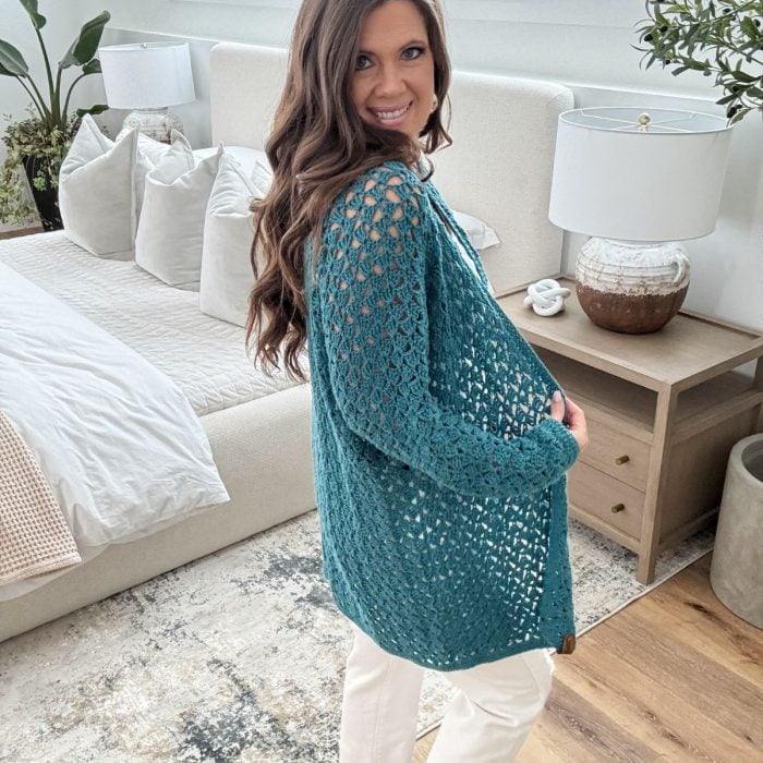 A person with long hair stands in a bedroom wearing a teal open-knit ruana and white pants, smiling at the camera. They are touching their ruana-covered midsection.