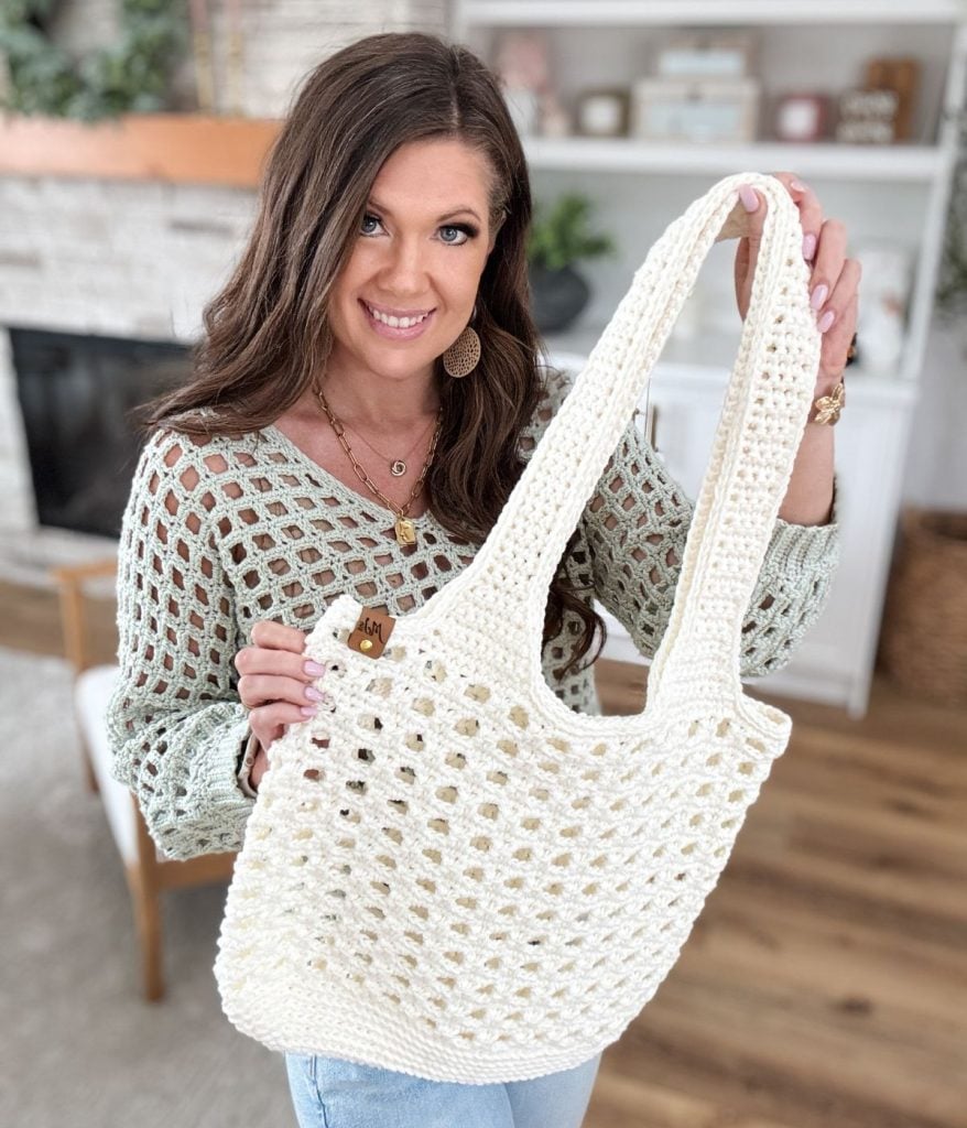 A woman stands in a cozy living room by the fireplace, holding a crocheted mesh bag. Her outfit—a matching crochet top—complements the bag beautifully, with shelves lined with books and trinkets providing a charming backdrop.