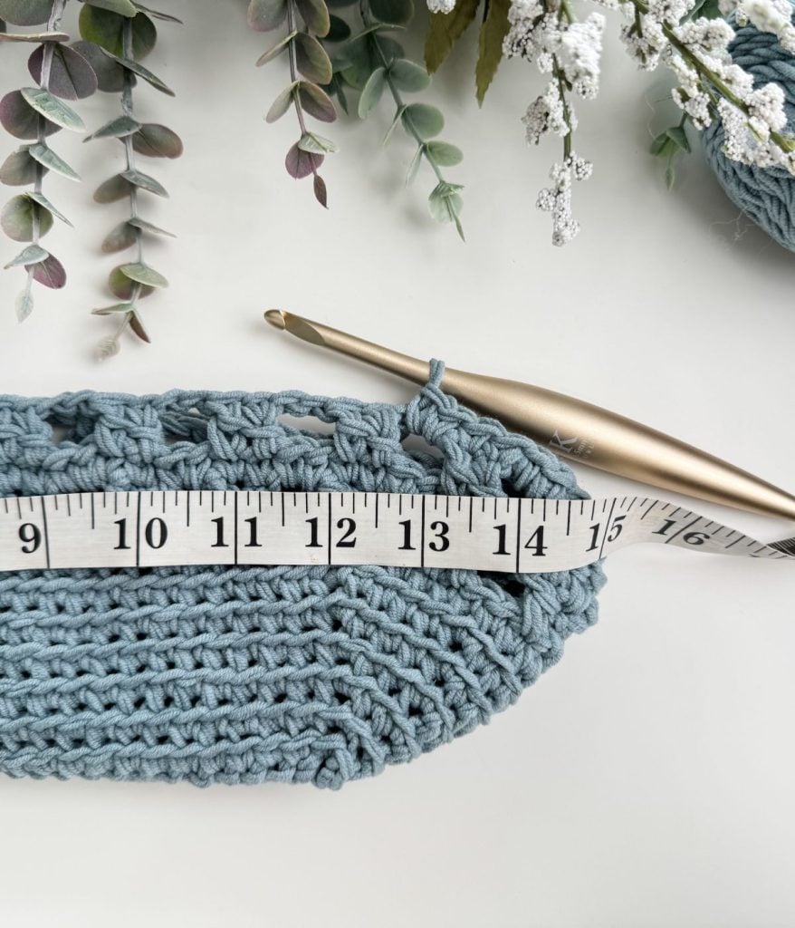 A crochet mesh bag takes shape with a gold crochet hook and a measuring tape marking 14 inches. Greenery adds a natural backdrop.
