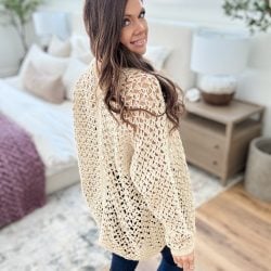 A person stands in a bright room, draped in a beige knitted ruana over their jeans, looking over their shoulder. Bedroom furniture and decor are visible in the background.