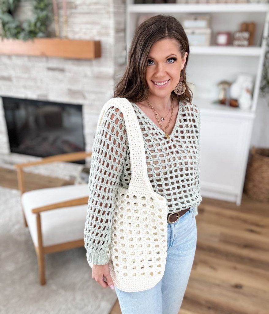 A person wearing a knitted sweater and jeans holds a stylish crochet mesh bag, standing in a living room with a fireplace and shelves in the background.