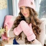 Woman wearing a pink knit hat with a fur pom-pom and matching mittens, standing indoors; text reads: "Heavenly Herringbone Hat Crochet Pattern & Video." Pairs perfectly with the Snowy Season Sweater for cozy winter style.
