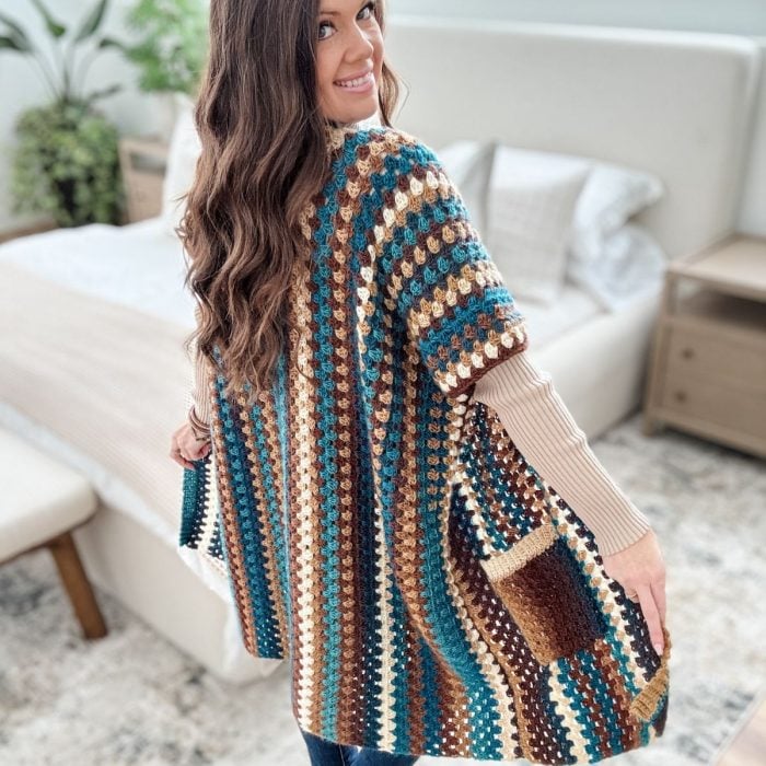 A person wearing a multicolored striped crochet cardigan, reminiscent of an easy free crochet pattern, stands in a bedroom with beige bedding and a nightstand.