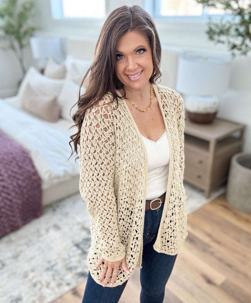 A woman in a beige crochet cardigan, reminiscent of a stylish ruana, and white top stands in a bright room with wooden flooring and neutral decor, smiling warmly at the camera.