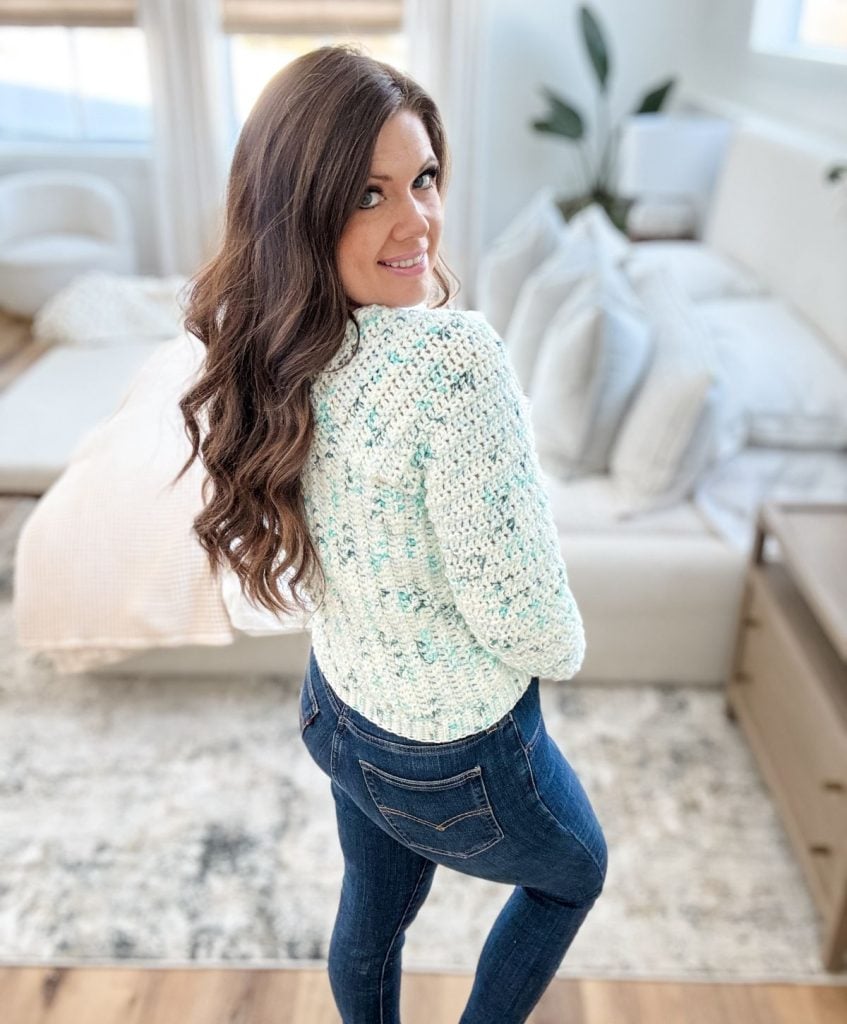 In a cozy living room, a person with long, wavy hair wearing a Snowy Season Sweater and jeans turns to look back at the camera, effortlessly showcasing the charm of this free crochet pattern.