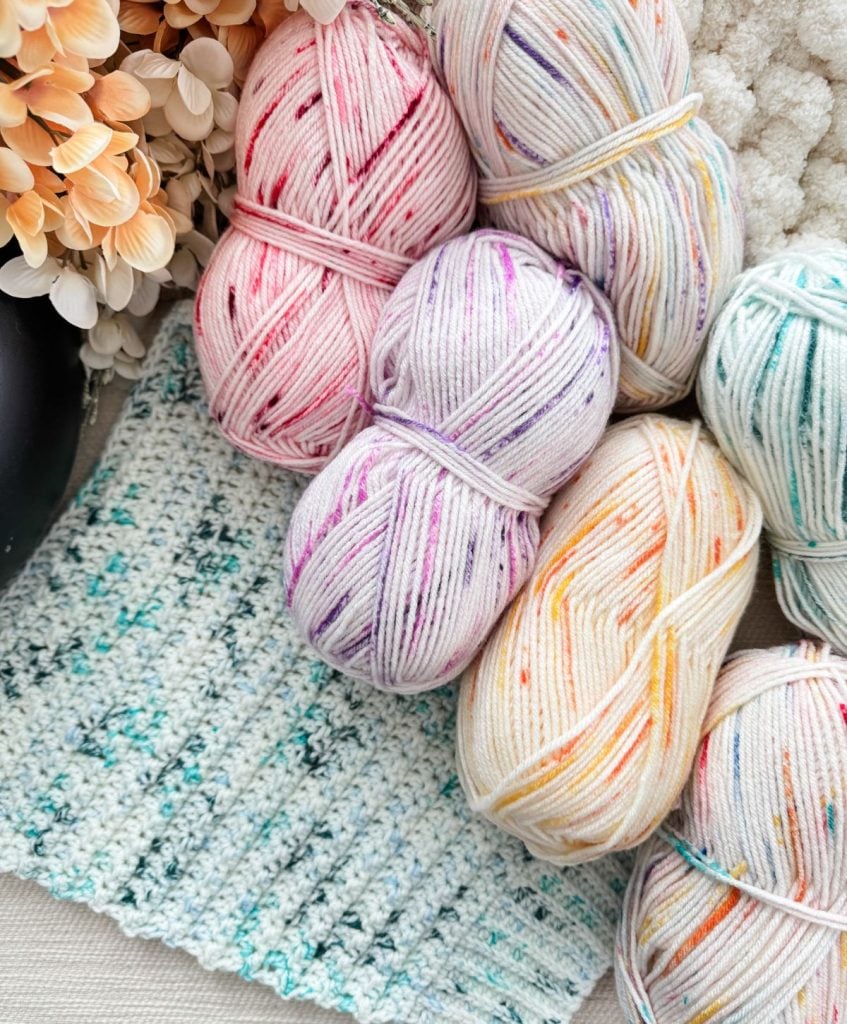 Colorful skeins of yarn in pink, purple, and orange tones sit beside a knitted swatch on a textured surface, poised for your next snowy season sweater project. Delicate flowers provide backdrop inspiration for your cozy crochet pullover pattern.
