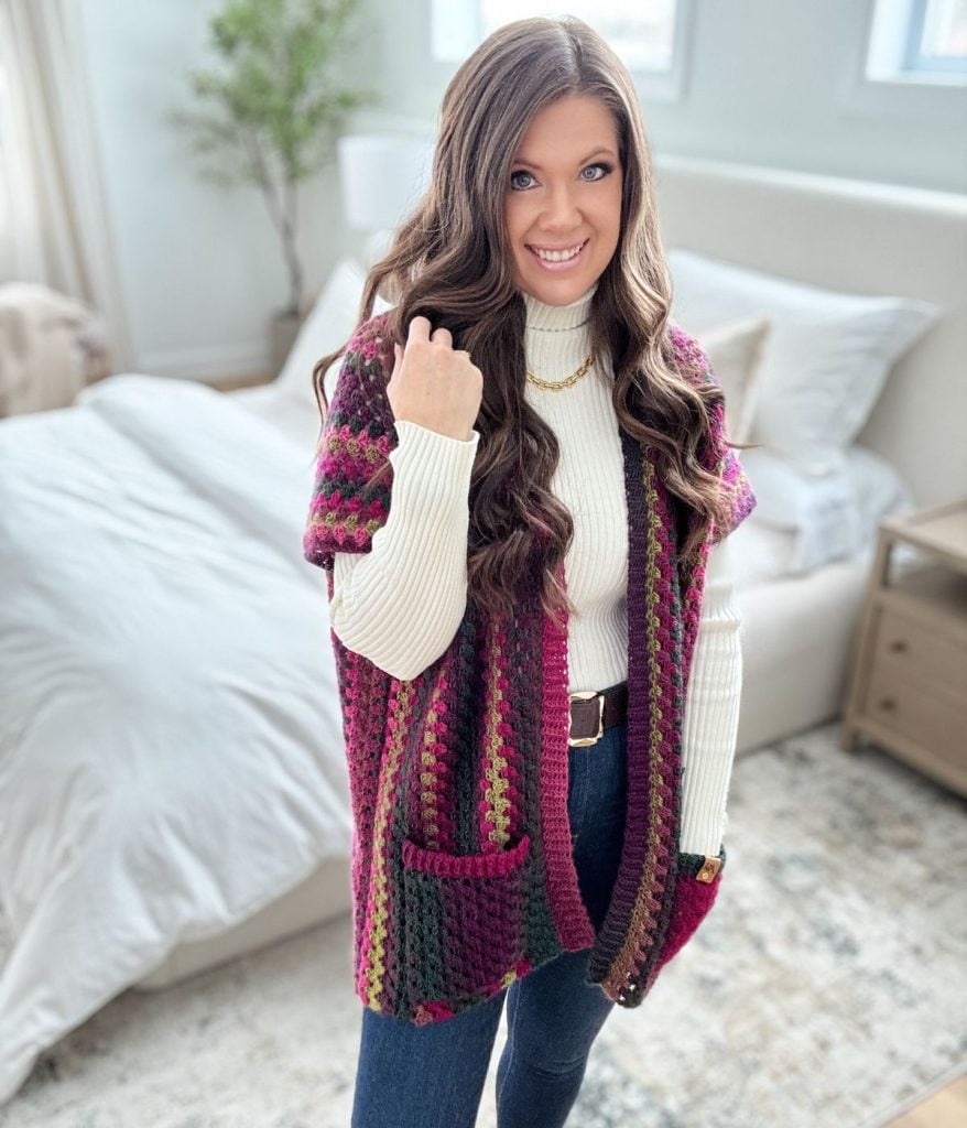 Woman with long hair wearing a crocheted vest over a white sweater stands in a bedroom, smiling. She has one hand on her hip, effortlessly flaunting her Granny Pop Ruana. A bed with pillows is visible in the background.