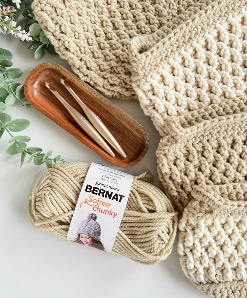 Beige crocheted fabric, two crochet hooks in a wooden tray, a skein of Bernat Softee Chunky yarn, a crochet basket, and green foliage rest on a white surface.