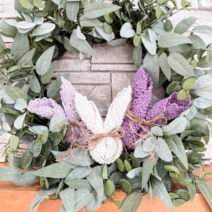 A leafy green wreath with three pairs of crocheted bunny ears in cream and purple, tied with twine bows and accented by a delicate crochet dishcloth, displayed against a stone background.