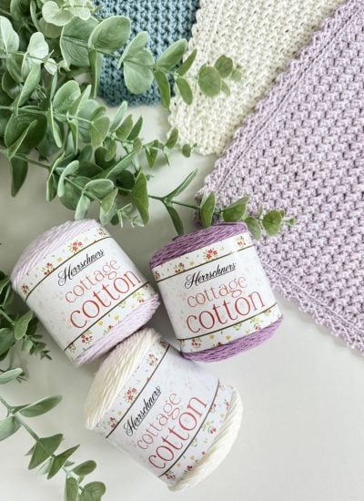 Three skeins of Herschners Cottage Cotton yarn in pastel colors are displayed next to green foliage and textured crochet dishcloth swatches.