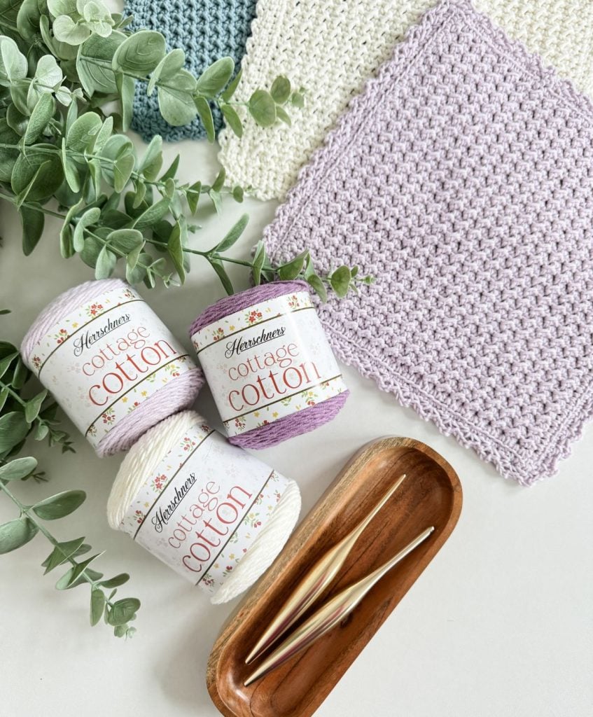 Three skeins of cottage cotton yarn, two crochet hooks in a wooden tray, two crochet dishcloth squares, and green leafy stems arranged on a white surface.