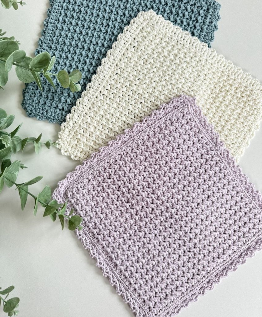 Three crochet dishcloths in blue, white, and light purple are neatly stacked on a light surface beside some green leafy stems.
