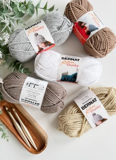 Five skeins of Bernat Softee Chunky yarn in neutral colors, with crochet hooks and a cozy crochet basket arranged on a wooden tray, green foliage partially visible in the corner.