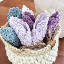 A cream-colored woven basket holds several folded crochet dishcloths in shades of blue, lavender, and purple, placed on a wooden surface.