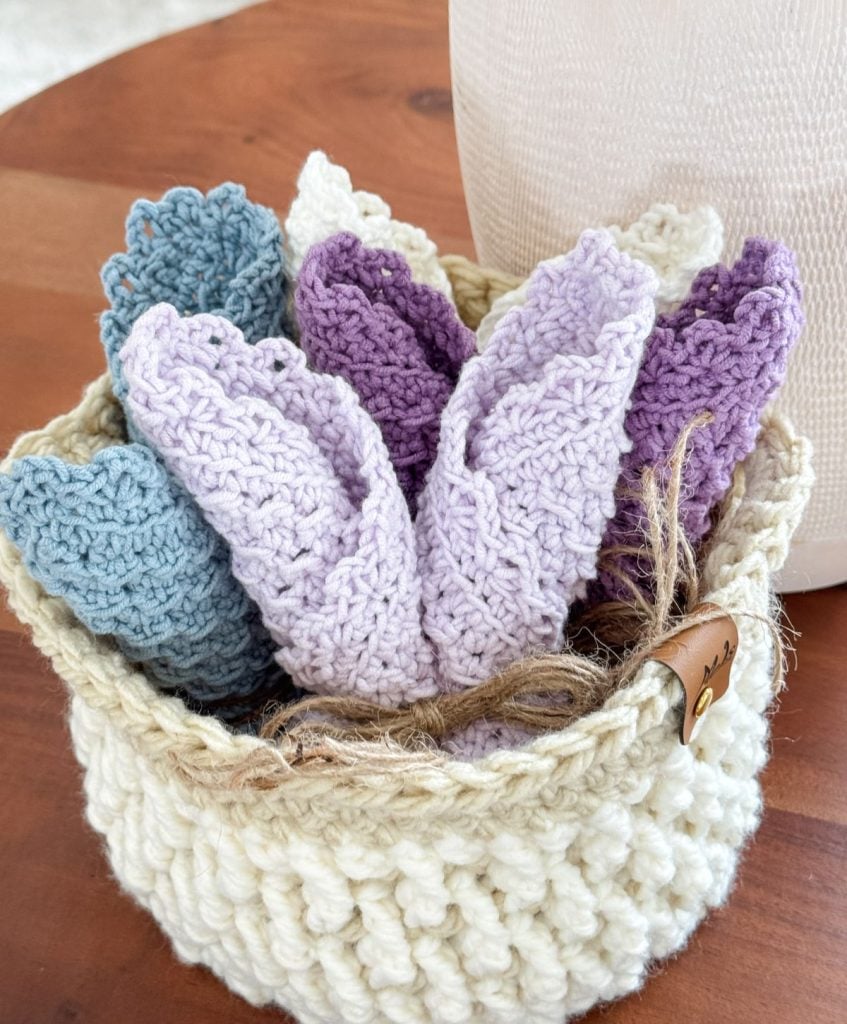 A cream-colored woven basket holds several folded crochet dishcloths in shades of blue, lavender, and purple, placed on a wooden surface.
