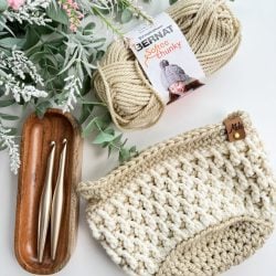 Crochet Basket, two aluminum crochet hooks in a wooden tray, a skein of Bernat Softee Chunky yarn, and artificial greenery are artfully displayed on a white surface.