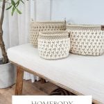 Three cream-colored crochet baskets in varying sizes sit on a cushioned wooden bench near a potted plant and window. Text reads: "Homebody Crochet Basket Pattern & Video - Bulky & Quick.