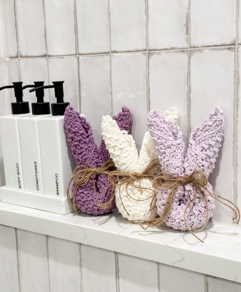 Three crocheted bunny-shaped decorations with twine bows sit on a bathroom shelf next to white bottles labeled body wash, conditioner, and shampoo. A neatly folded crochet dishcloth adds a cozy touch against the white tiled wall in the background.