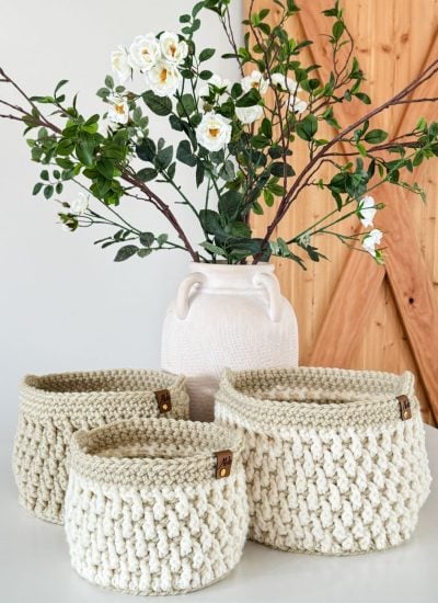 A white vase with white flowers and green leaves stands behind three cream-colored crochet baskets on a white surface.